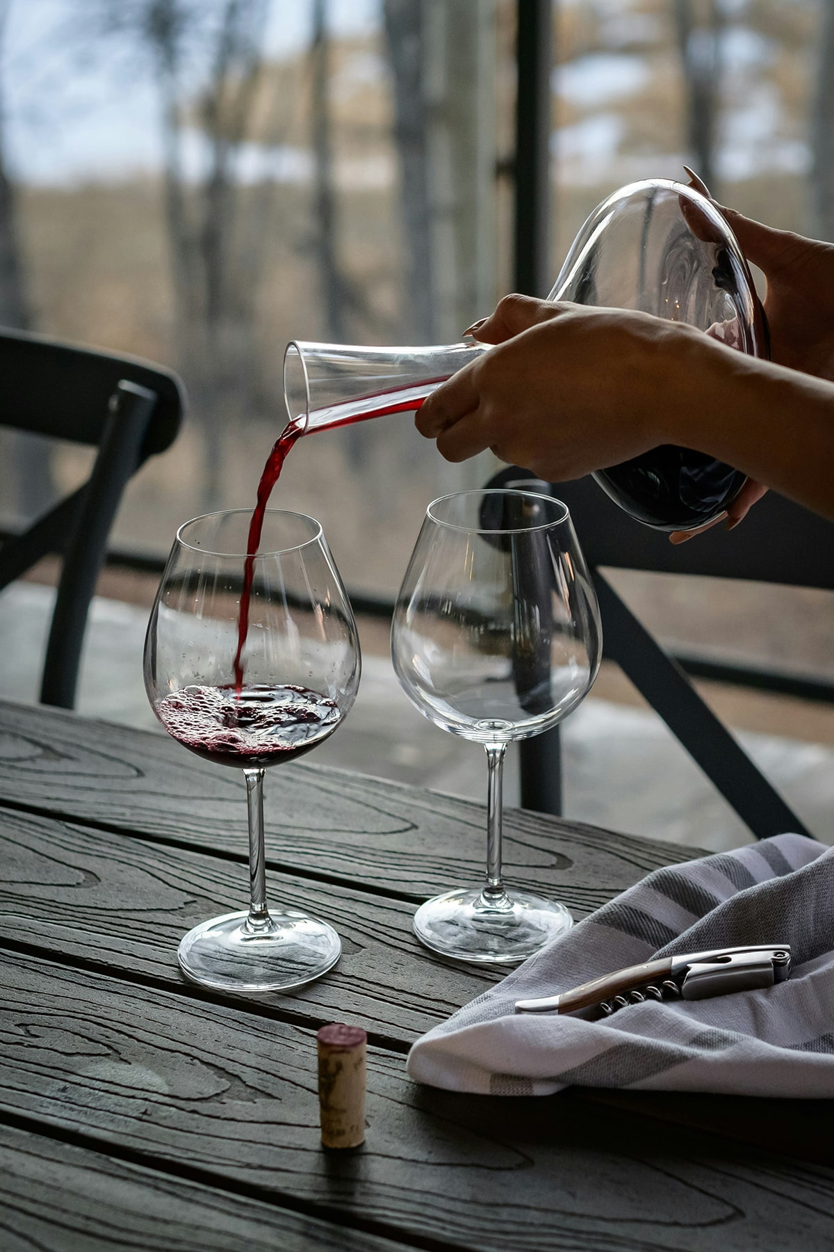 Rode wijn dat ingeschonken wordt door een vrouw op een houten eettafel met vanuit het raam uitzicht op een bos met herfstkleuren